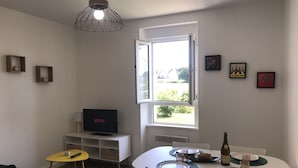 A modern living room with a TV, dining table, and a window with an open door. - Renovated one-bedroom apartment, 300 m from the beach, free private parking (Bénodet)