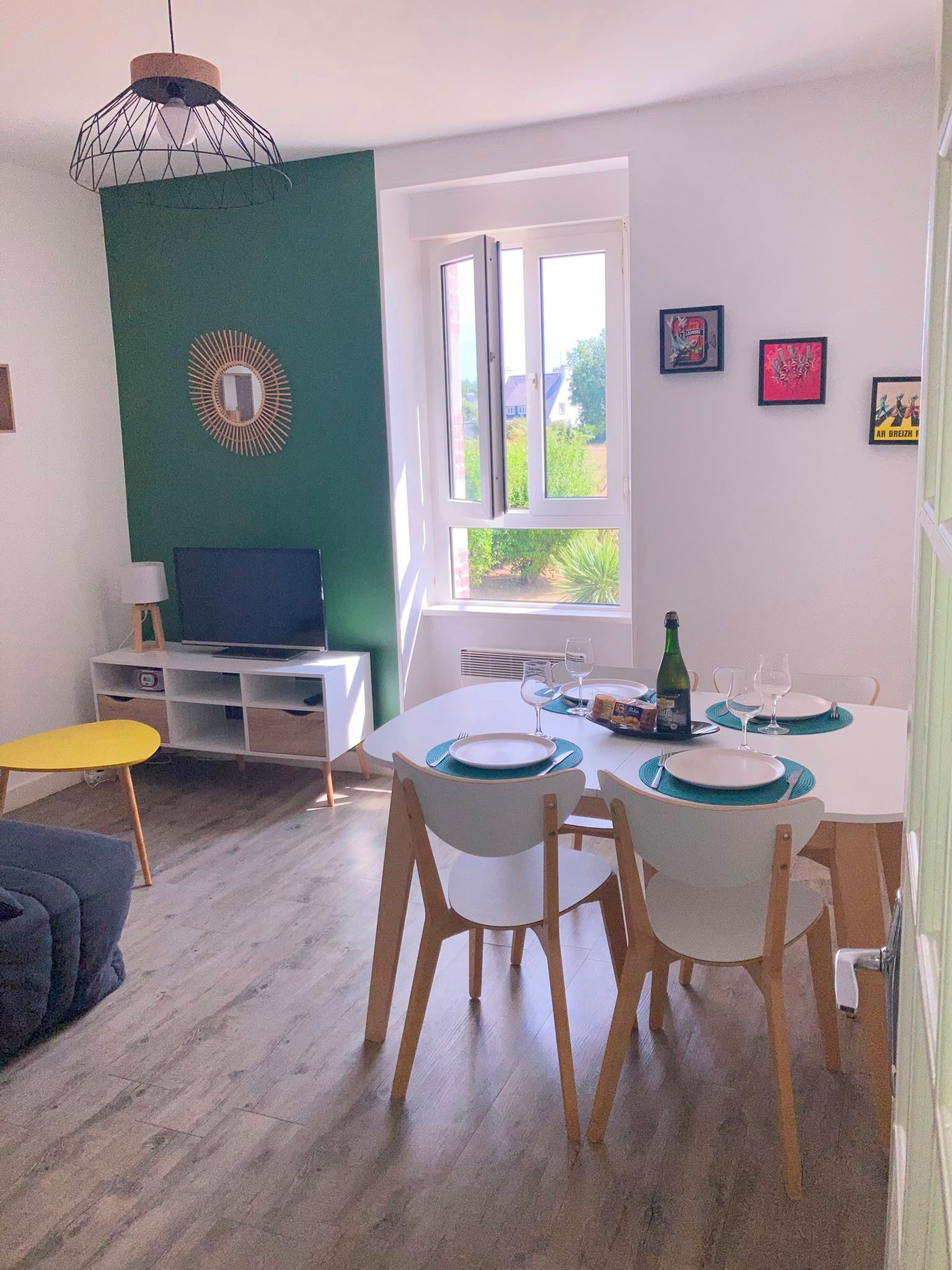 Un coin repas avec une table dressée pour quatre personnes, un téléviseur posé sur un meuble et une fenêtre donnant sur un paysage verdoyant.