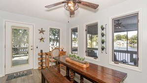 Ein Essbereich mit Holztisch, Bänken und Deckenventilator. Fenster bieten Blick auf eine Terrasse und einen Außenbereich.