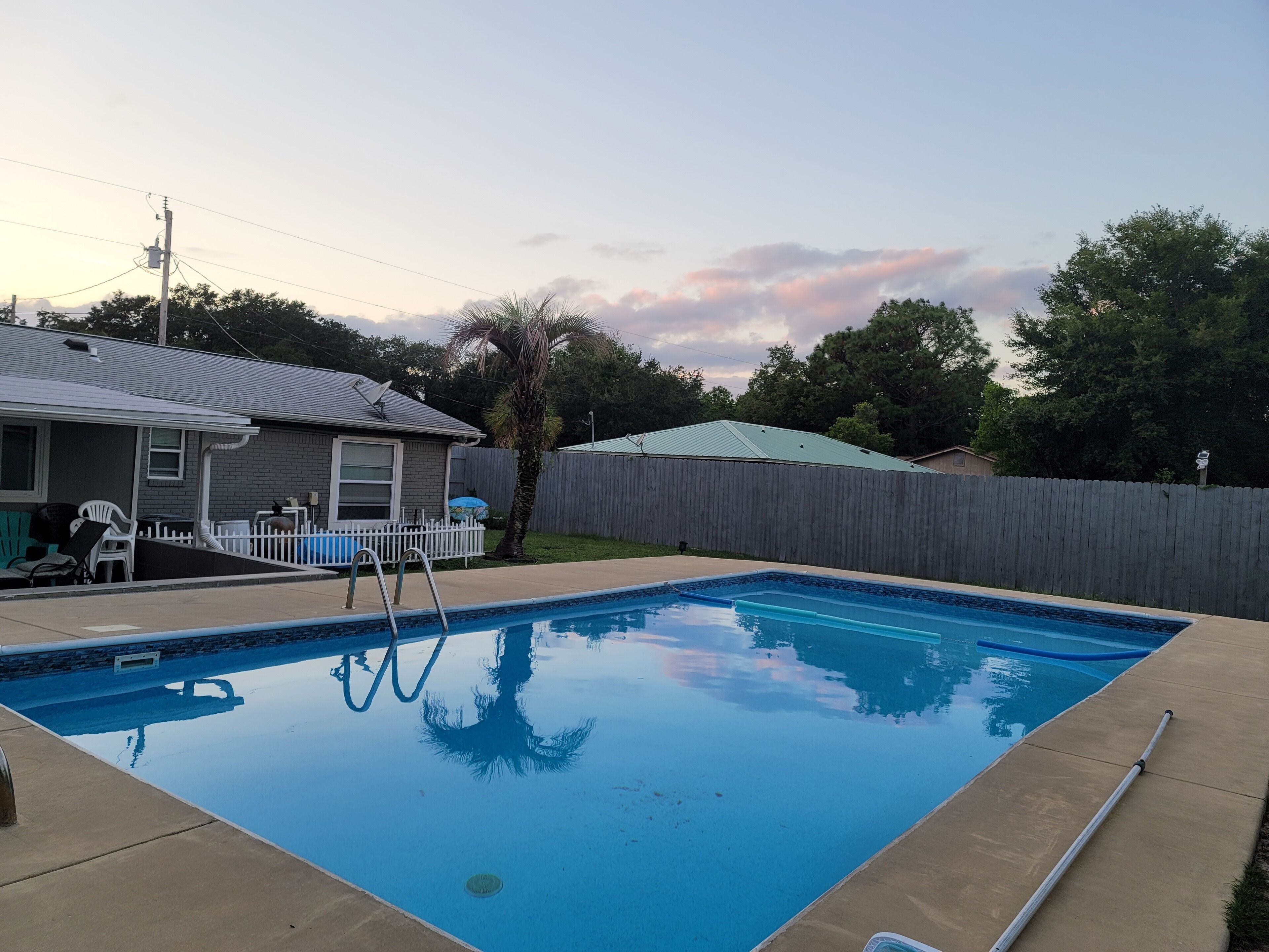 A rectangular swimming pool with a ladder, surrounded by a wooden fence, and a house with a satellite dish in the background.