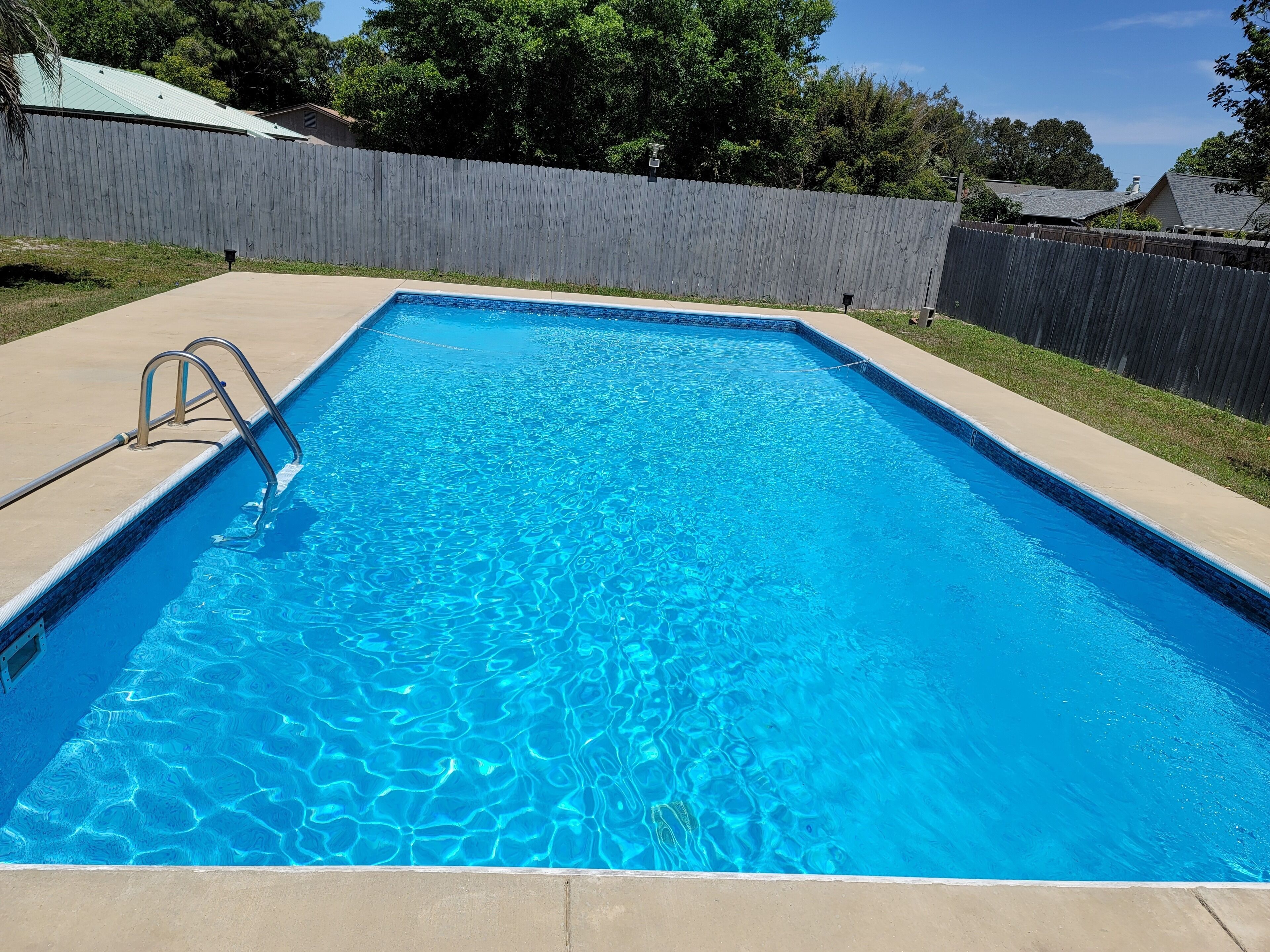 A rectangular swimming pool with clear blue water, surrounded by a concrete deck and a wooden fence.
