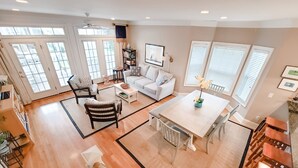 Un vaste espace de vie comprenant un canapé blanc, une table basse et une table à manger avec des chaises. De grandes fenêtres équipées de stores, un ventilateur de plafond et un escalier menant à un autre niveau complètent l'ensemble.