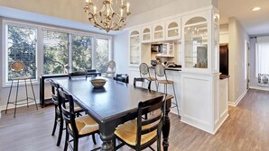 Un comedor con una gran mesa de madera, seis sillas y una araña. Hay ventanas amplias con persianas, una zona de cocina con armarios blancos, y vistas a árboles afuera.