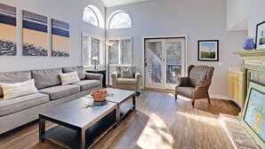 Un salón amplio con un sofá gris, un sillón estampado y una mesa de centro. Hay ventanas grandes y un ventanal corredero que da a un área exterior.