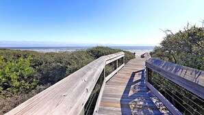 Una pasarela de madera que conduce a una playa, con vistas despejadas al océano y montañas distantes.