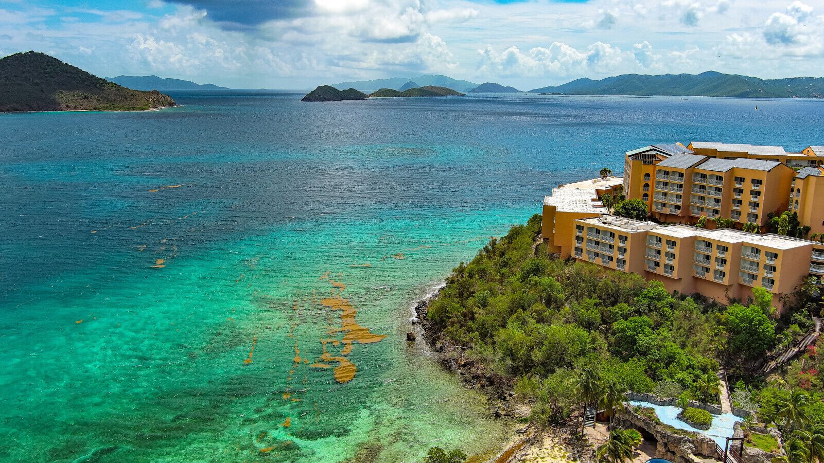 A coastal hotel with multiple buildings, a clear view of the turquoise sea, and a rocky shoreline with greenery.