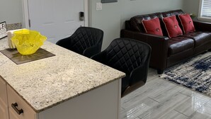 A modern kitchen with a granite countertop, black chairs, a brown leather sofa with red cushions, and a blue and white rug.