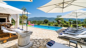 Un área junto a la piscina con sillones reclinables, sombrillas y piso de baldosas de piedra.