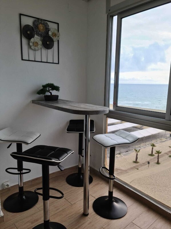 A corner room with a large window overlooking a beach, a small potted plant, and three bar stools.