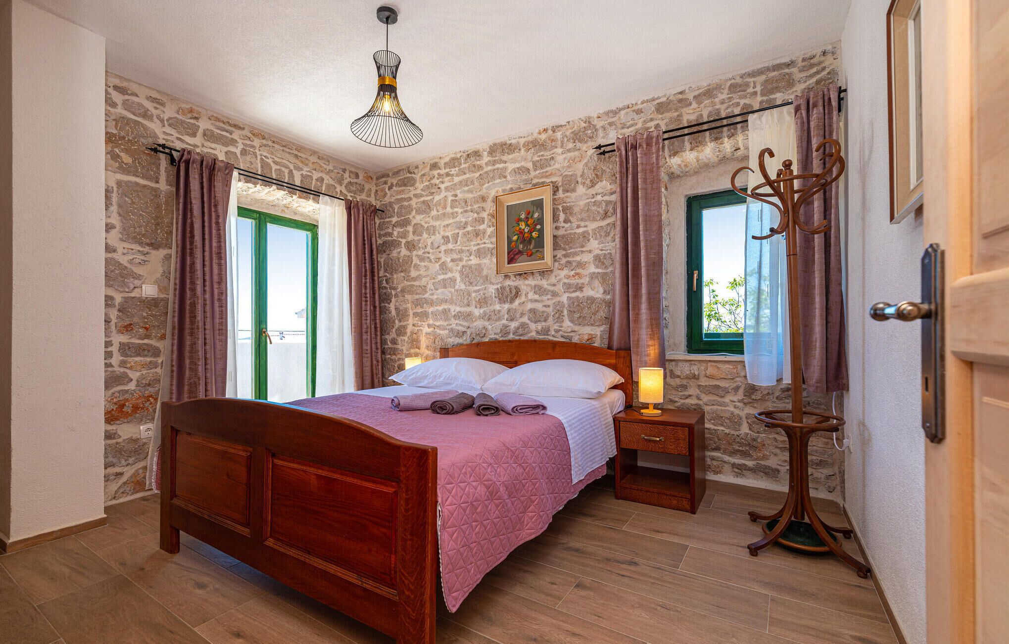 Una camera da letto con parete in pietra, un letto di legno, un comodino, una lampada, una finestra con tende e un appendiabiti.