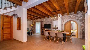 Een ruime woonkamer met een houten plafond, stenen muren, een eettafel met stoelen en een televisie.