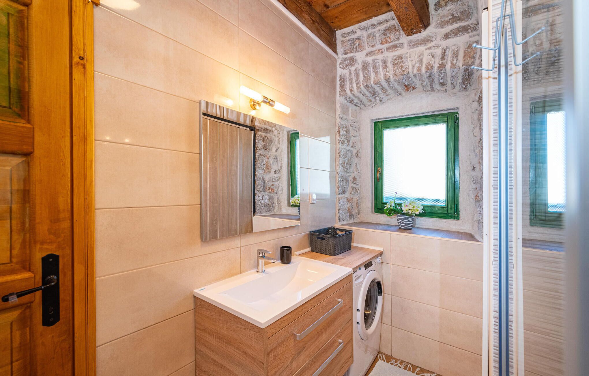 A bathroom with a wooden vanity, a mirror, a washing machine, and a window with green shutters.