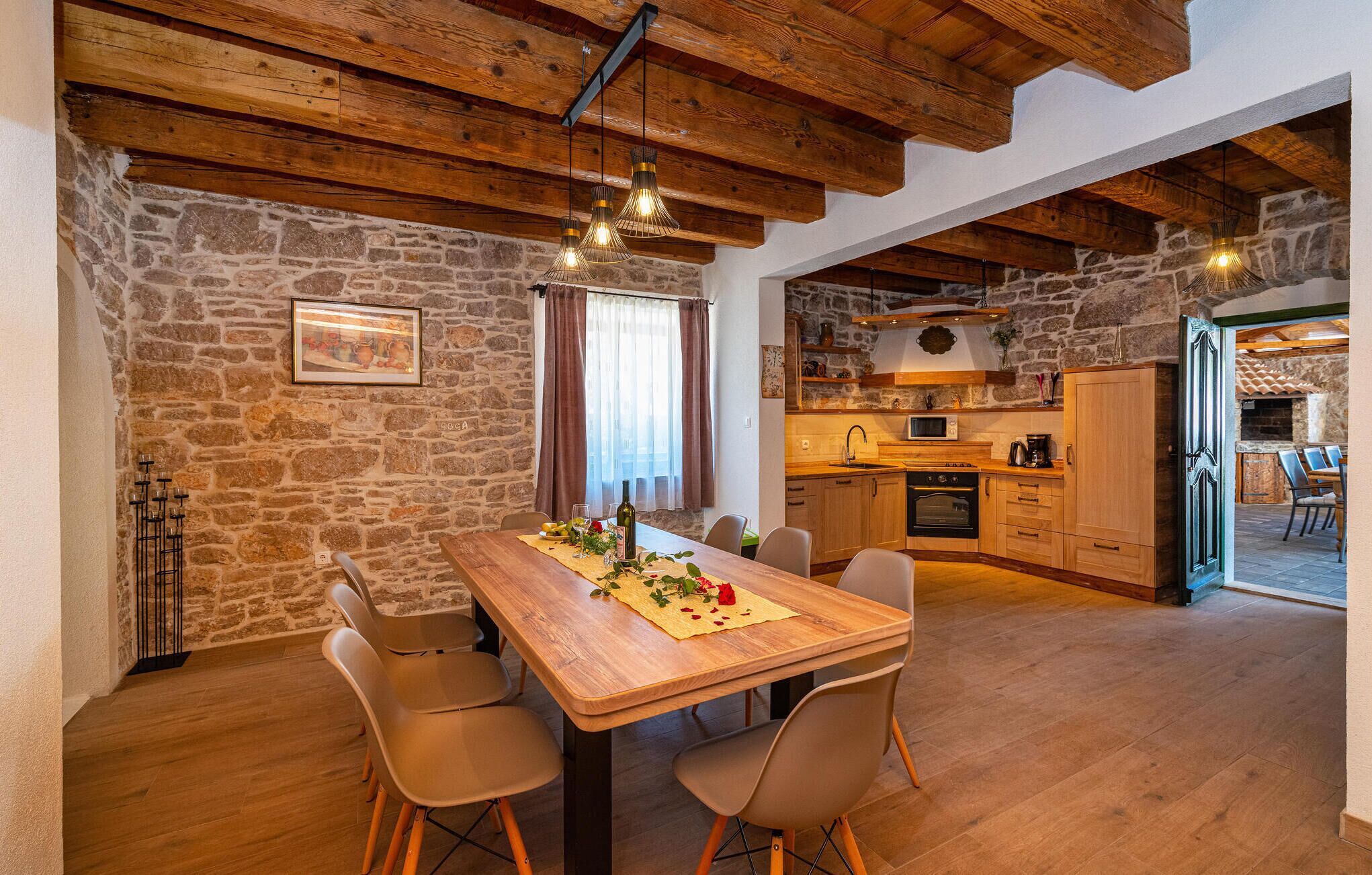 Un comedor rústico con una mesa de madera, cuatro sillas y una pared de piedra. Se ve una cocina al fondo con armarios de madera y una chimenea de piedra.