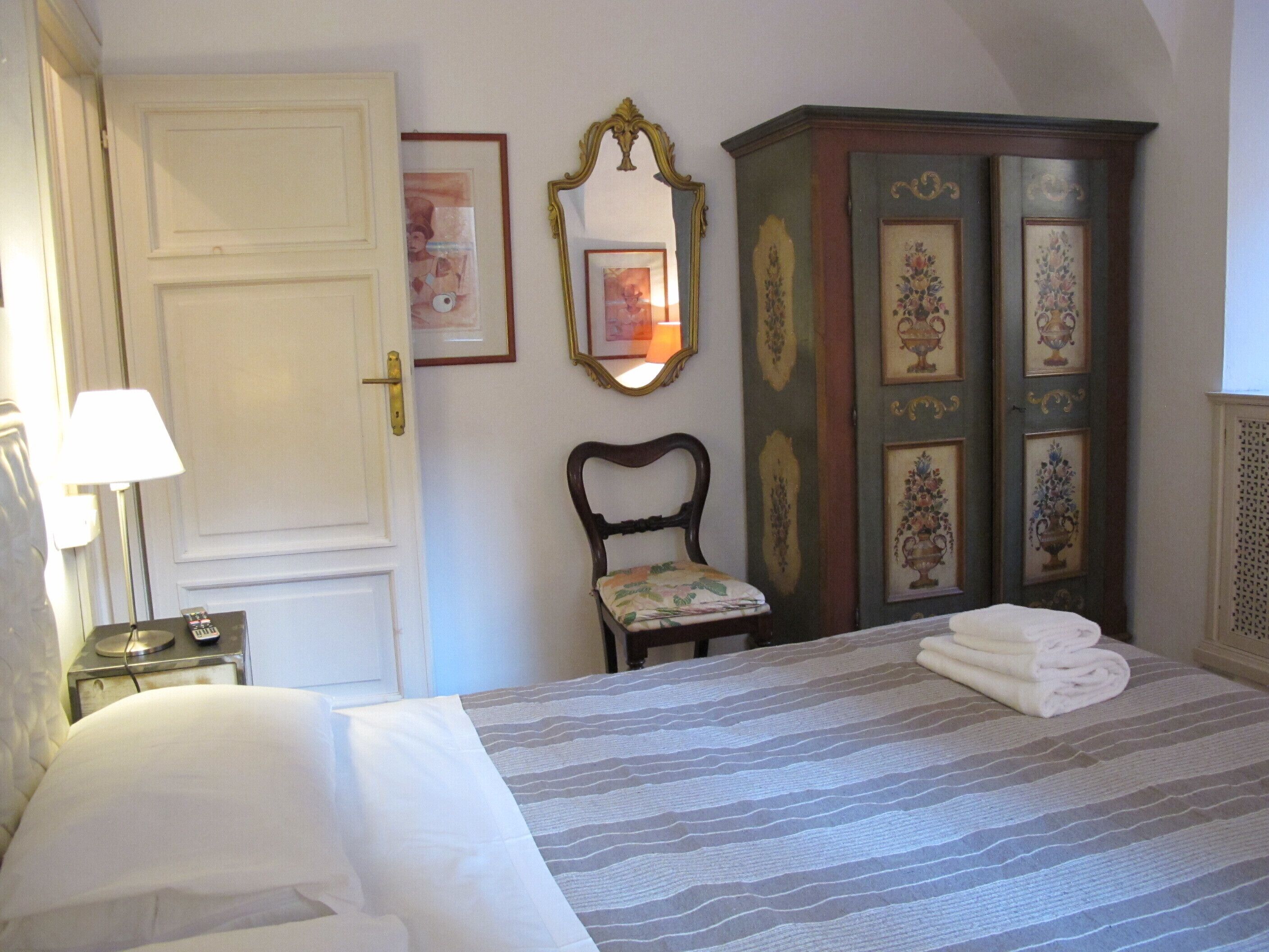 A hotel room with a bed, a chair, a wardrobe, and a bedside table with a lamp.