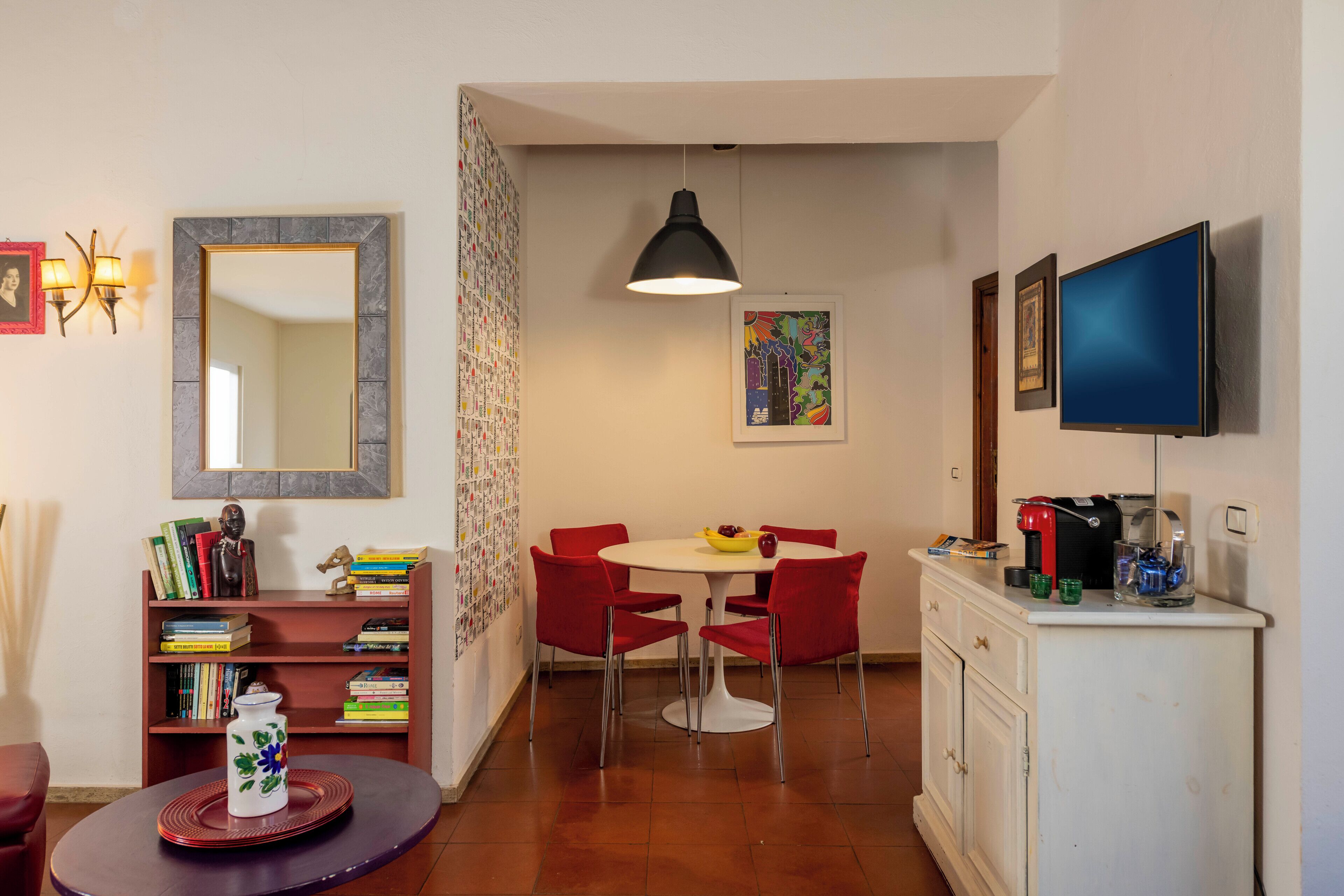 Een compacte woonruimte met een eettafel, rode stoelen, een witte kast, een televisie en een boekenkast.