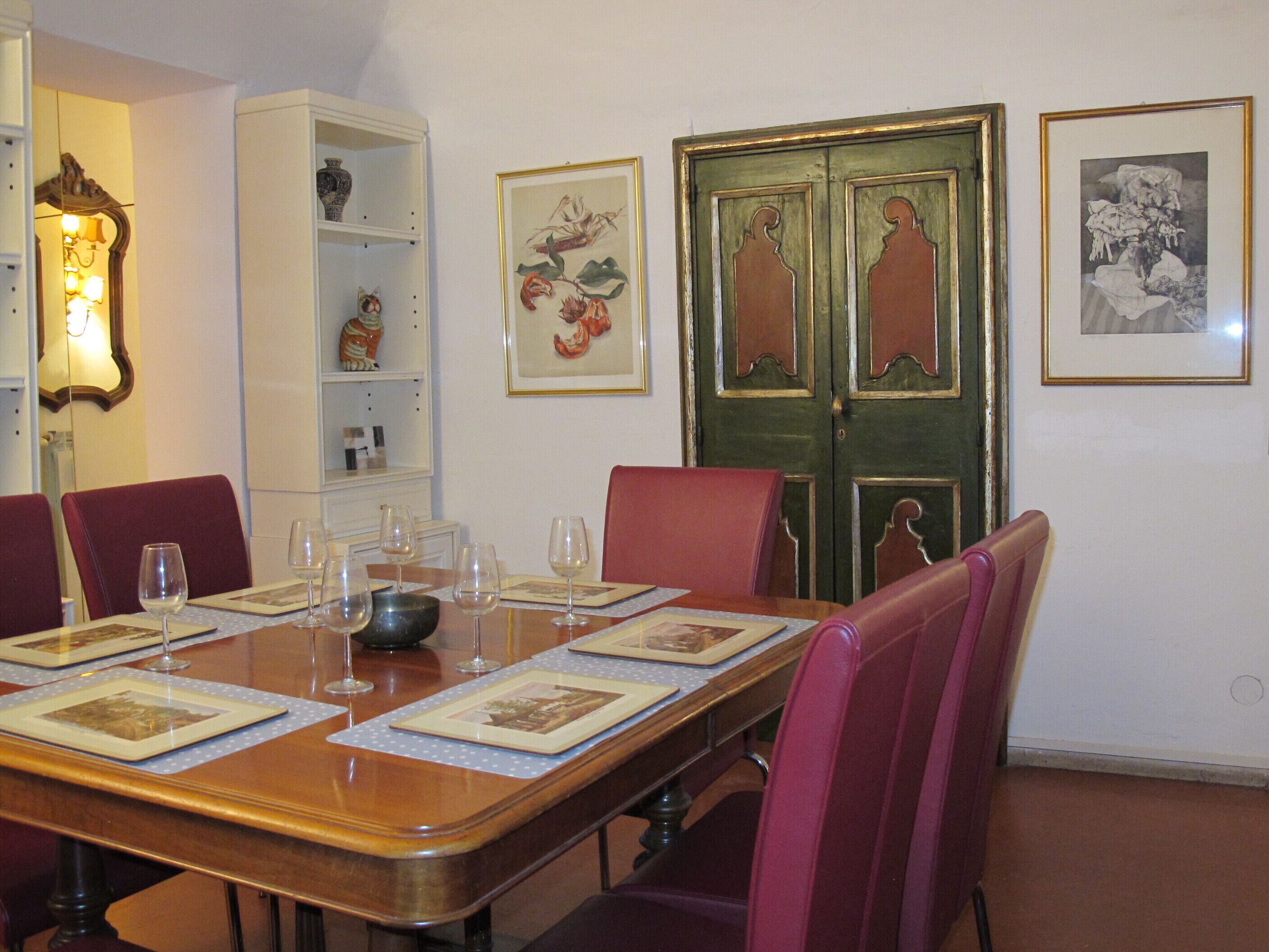 A dining room with a wooden table set for six, surrounded by red chairs. There are framed botanical prints on the wall and a green door with decorative panels.