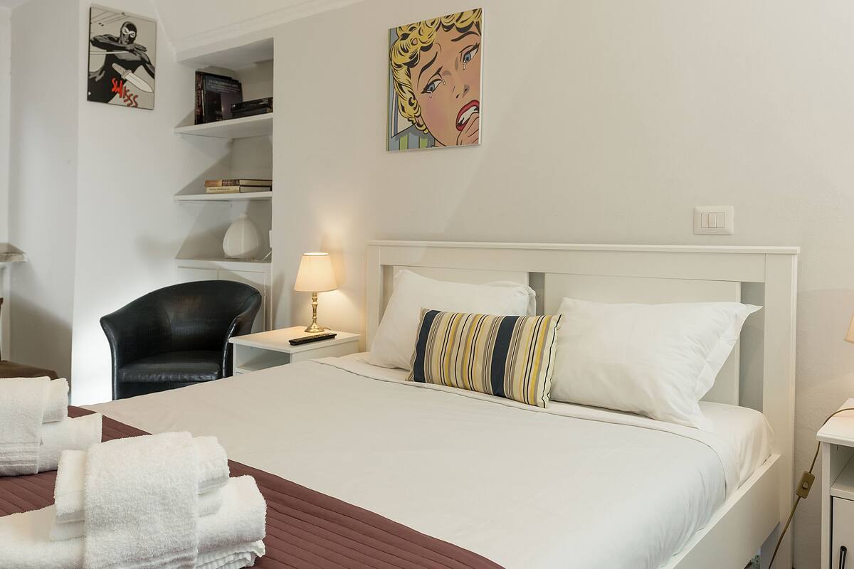 A hotel room with a bed, two bedside tables, a chair, a lamp, a shelf with books, and a painting on the wall.