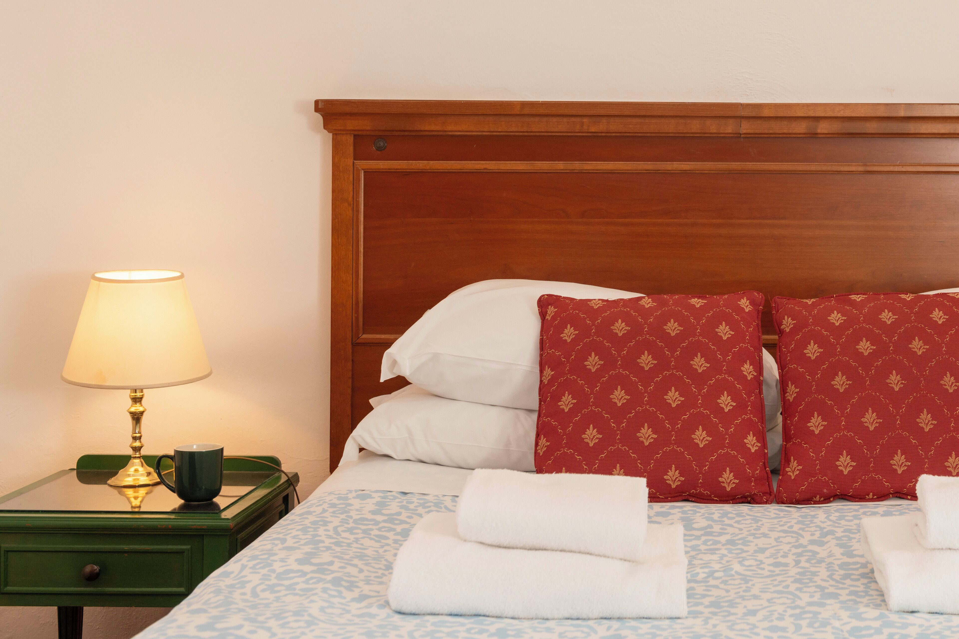 A neatly made bed with white and red pillows, a green nightstand with a lamp, and a blue patterned bedspread.