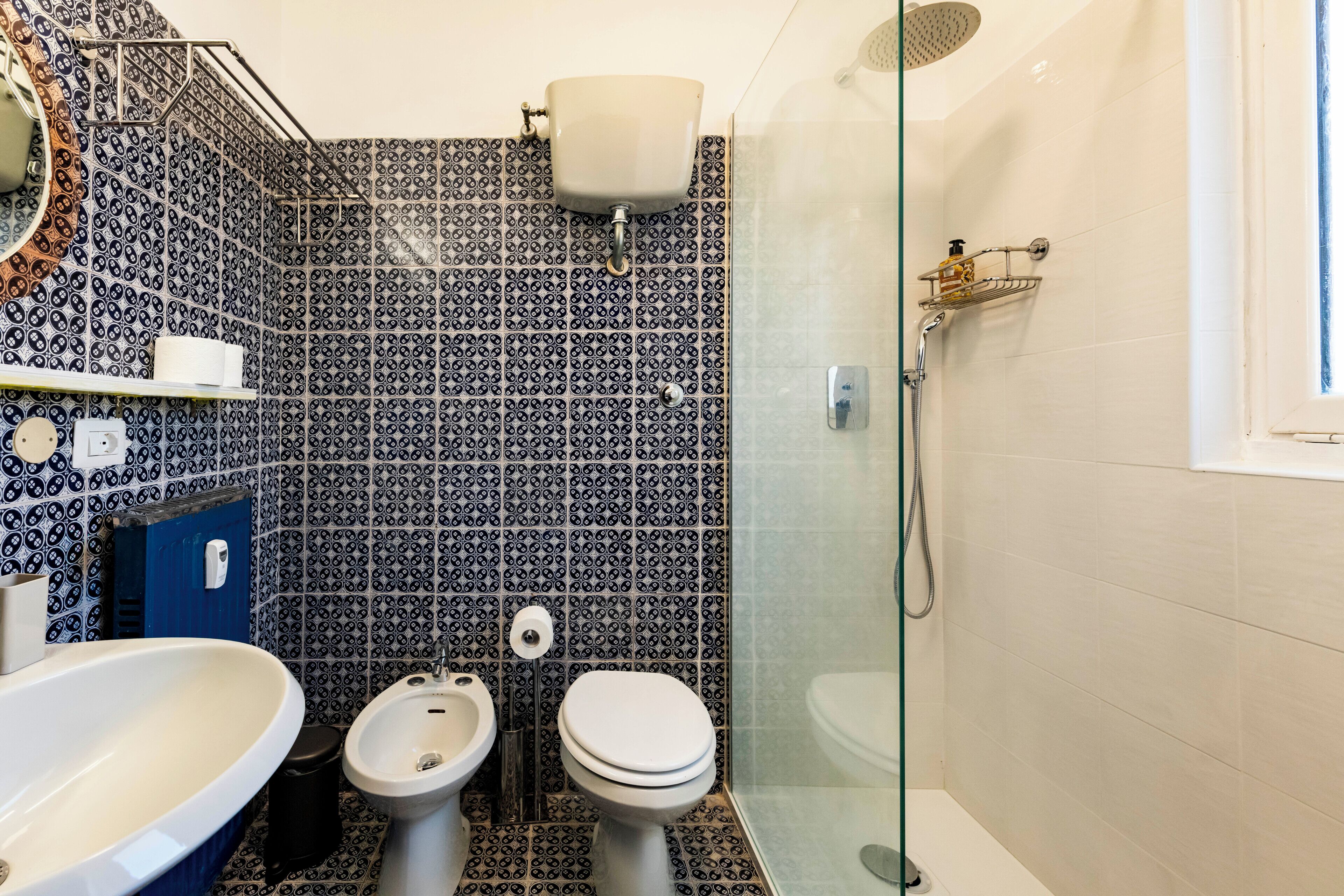 A bathroom with a patterned tile wall, a sink, two toilets, a bidet, and a glass-enclosed shower.