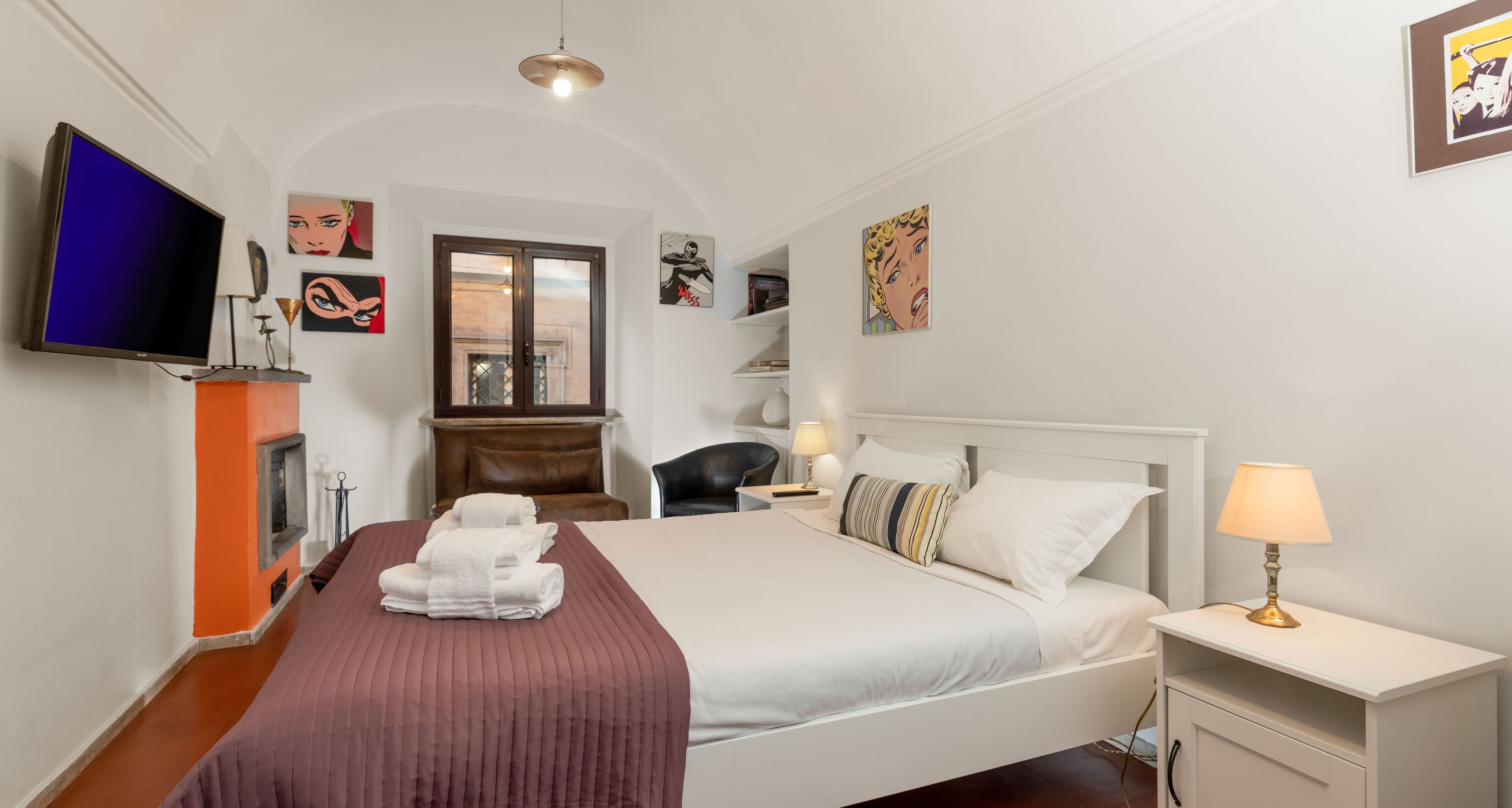A hotel room with a bed, a TV, a fireplace, and artwork on the walls.