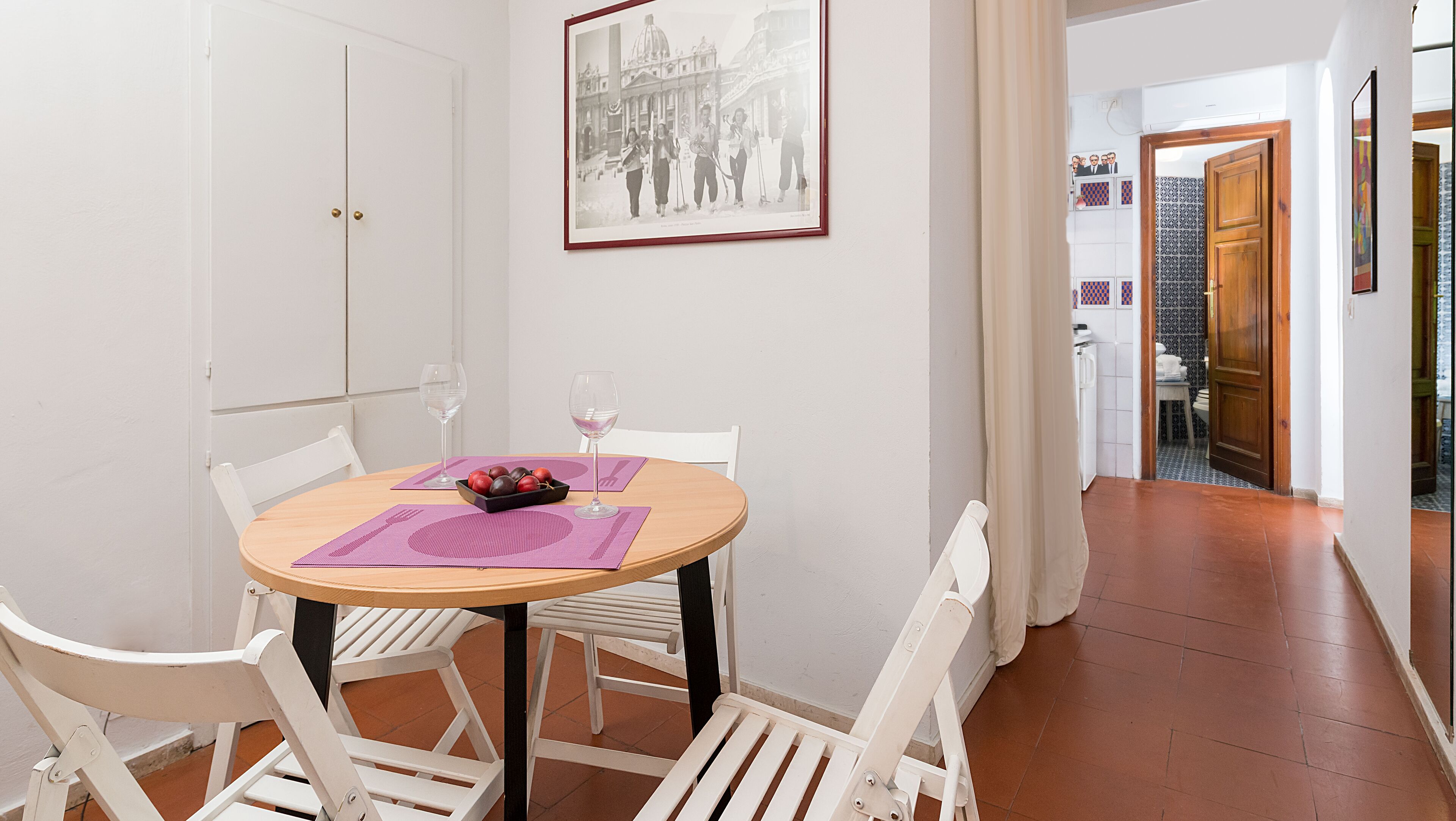 Un comedor con una mesa redonda, sillas blancas, un cuenco de fruta y copas de vino.