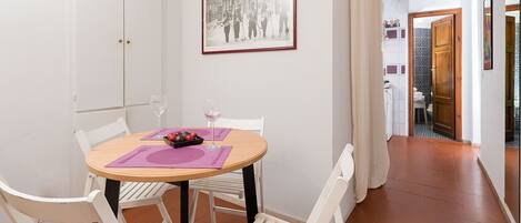 Un comedor con una mesa redonda, sillas blancas, un cuenco de fruta y copas de vino.
