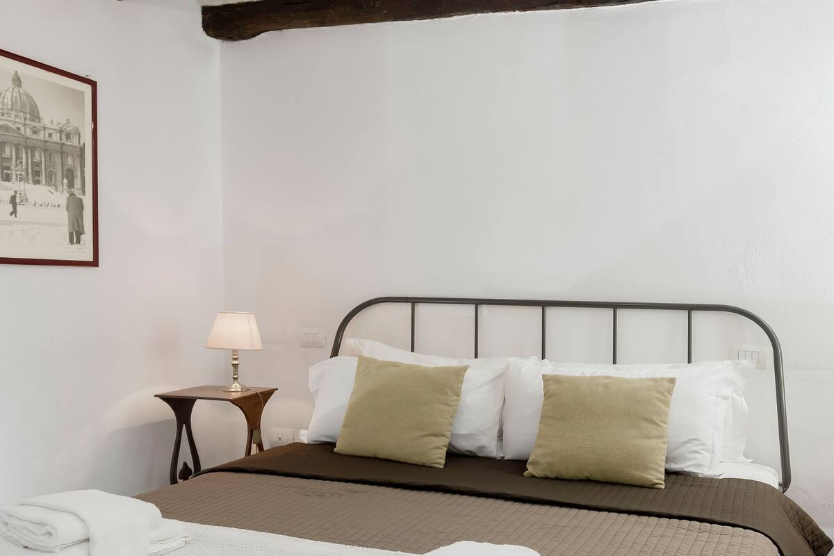 A neatly made bed with a metal headboard, a bedside lamp, and a framed picture of a historical building on the wall.