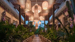 Un sentier en bois, bordé de plantes tropicales, mène à un bâtiment doté d’un toit de chaume et orné de lanternes suspendues.
