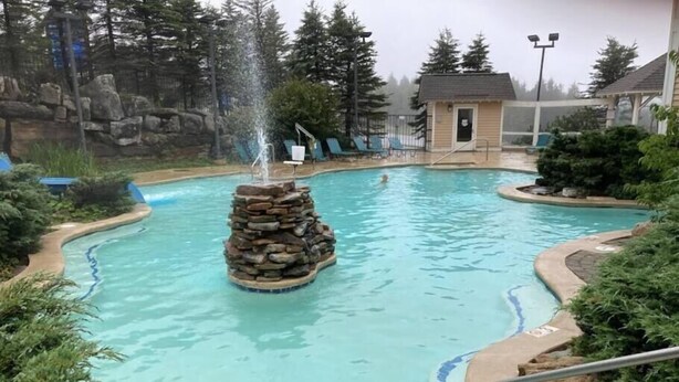Une piscine avec une fontaine en pierre, entourée d’arbres et d’un petit bâtiment.