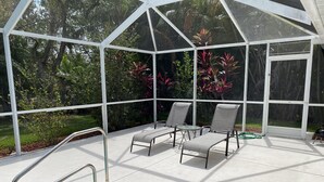 A covered outdoor pool area with two lounge chairs and a small table, surrounded by a white fence and lush greenery. - Beautiful Private Florida Pool Home (Cape Coral)