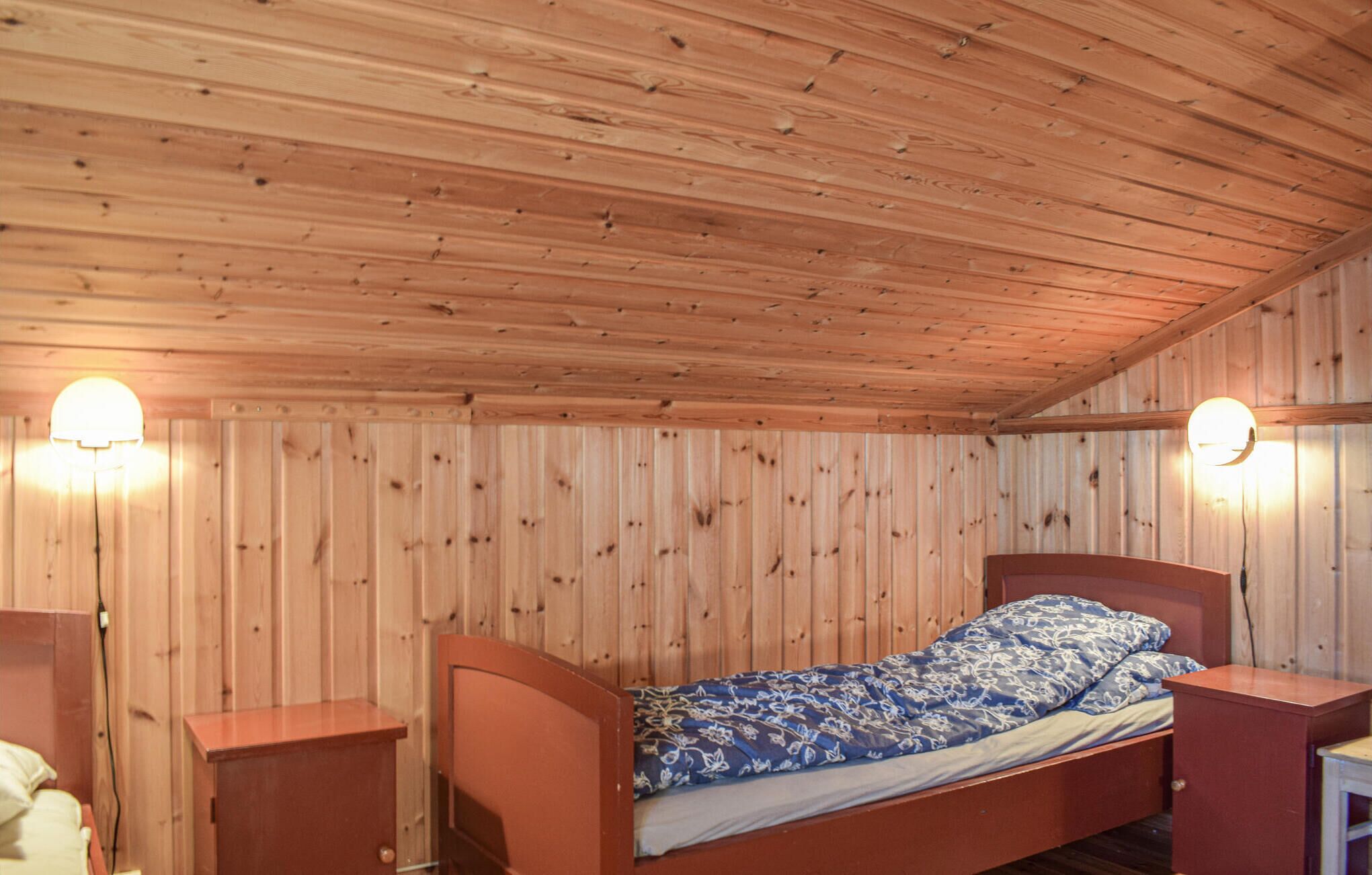 Un lit simple dans une chambre aux murs et au plafond en bois, avec deux lampes de chevet et une petite table de chevet en bois de chaque côté du lit.
