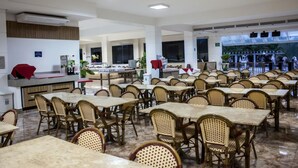 A spacious dining area with multiple tables and wicker chairs, a reception desk, and large windows offering a view of the outdoors. - Lindo Flat Localizado no Lacqua Diroma V (GO)