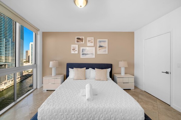 A hotel room with a large bed, two bedside tables, a nightstand, a view of a cityscape through large windows, and artwork on the wall.