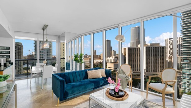 Ein geräumiges Wohnzimmer mit einem blauen Sofa, einem weißen Couchtisch und Korbstühlen. Große Fenster bieten Blick auf die Stadt.