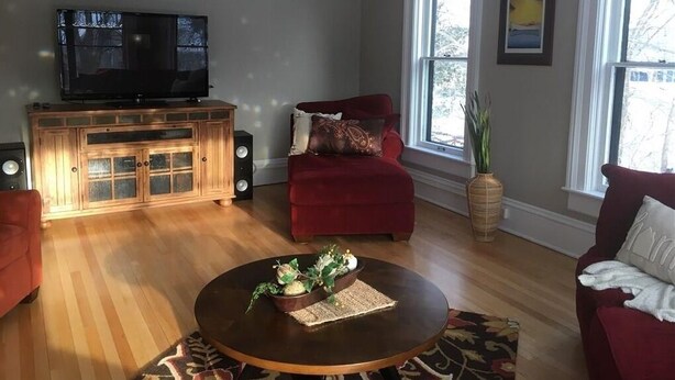 A living room with a wooden entertainment center, a red sofa, a round coffee table with decorative items, and large windows providing natural light. - Close to Downtown & UWEC (Eau Claire)