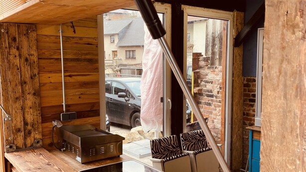 A rustic wooden interior with a wooden bench, a small table, and a window with a view of a parked car and houses outside. - Vintage Home 55 (Eisenach)