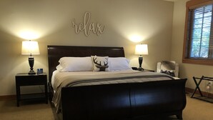 A bedroom with a dark wooden bed, two bedside lamps, a wall-mounted 'relax' sign, a window with blinds, and a small table with a lamp. - EV Charger! HOT TUB! Perfect family getaway in the ❤️ of Suncadia! (Cle Elum)