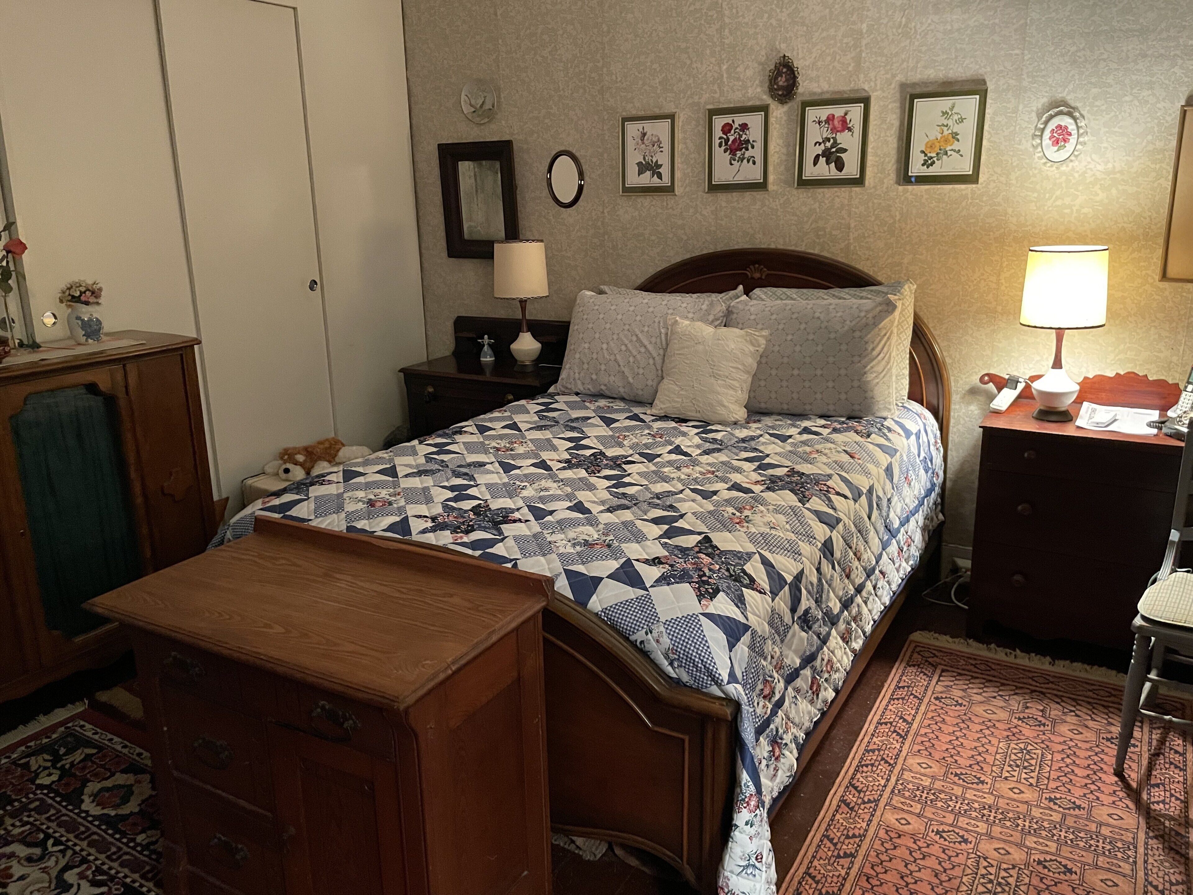 Un dormitorio con cama de madera, una mesita de noche con lámpara, cómoda, una alfombra pequeña y una pared con cuadros florales.