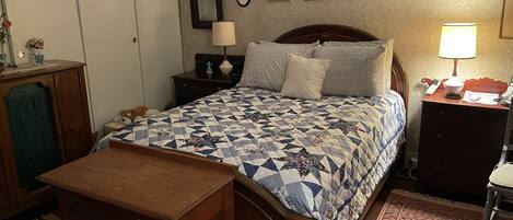 Un dormitorio con cama de madera, una mesita de noche con lámpara, cómoda, una alfombra pequeña y una pared con cuadros florales.