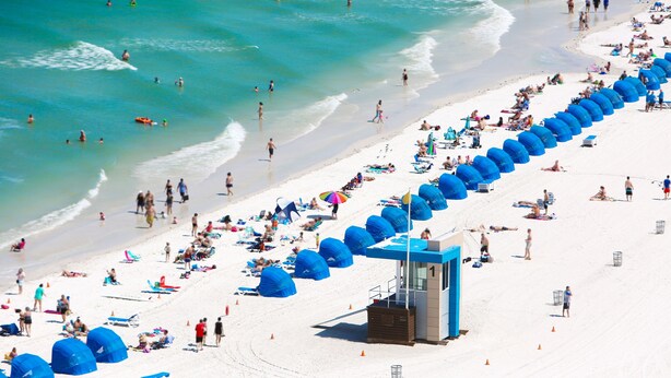 A beach with rows of blue umbrellas and beach chairs, a lifeguard tower, and people enjoying the beach.