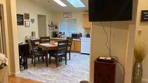 Ein Wohnzimmer mit grauem Sofa, einem Holztisch und einem Essbereich mit Tisch und Stühlen. Eine Deckenlampe, ein flachbildfernseher an der Wand und eine Küche im Hintergrund sind ebenfalls zu sehen.