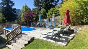 Ein Garten mit Swimmingpool, Holzterrasse, Liegestühlen und einem roten Sonnenschirm.