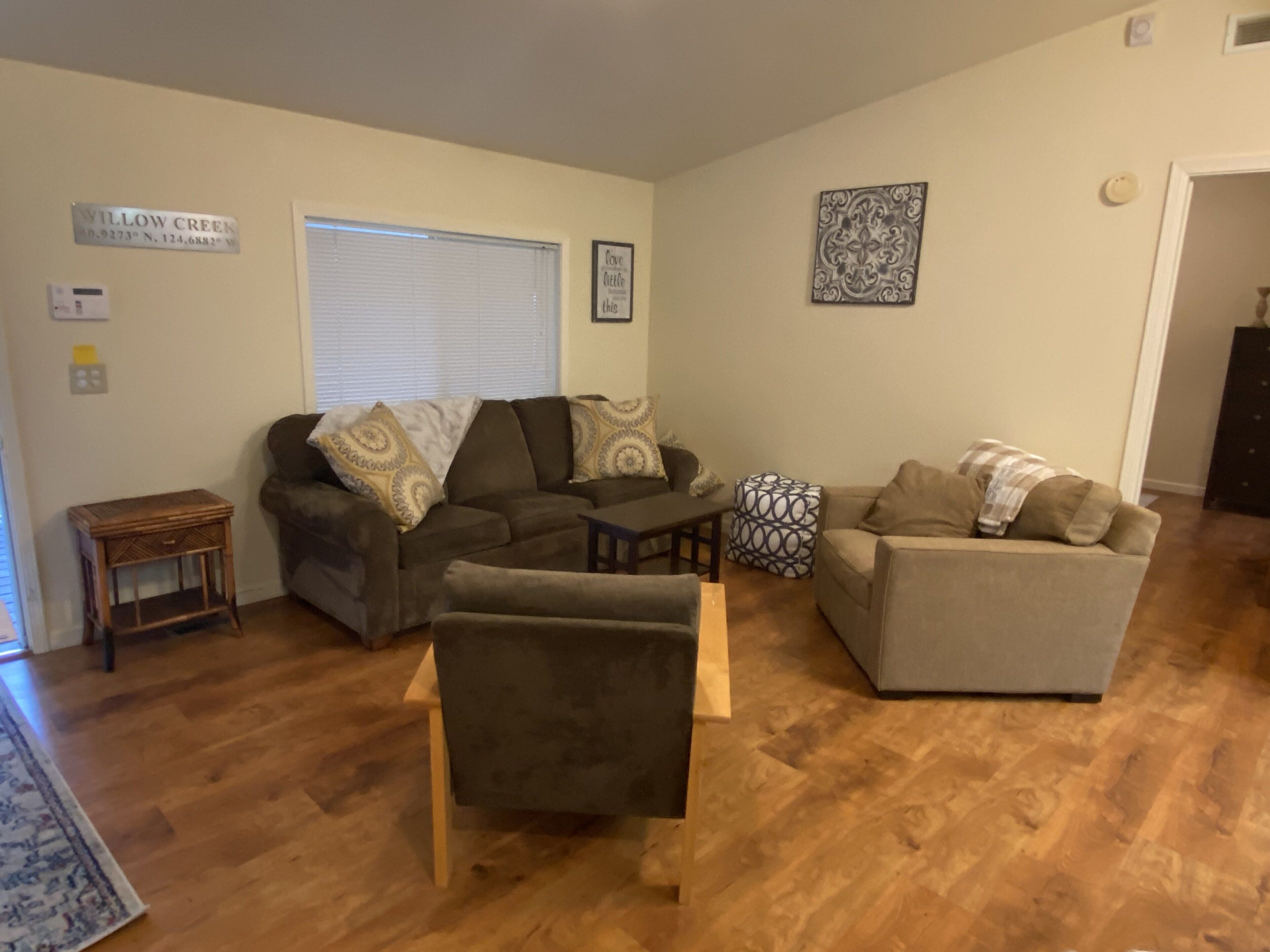 A living room with a brown sofa, a grey sofa, and a wooden coffee table. There is a window with blinds, a small wooden side table, and a decorative wall piece.