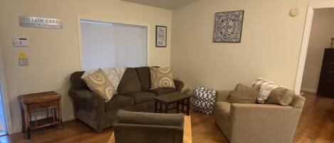 A living room with a brown sofa, a grey sofa, and a wooden coffee table. There is a window with blinds, a small wooden side table, and a decorative wall piece.