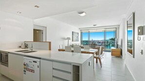 A modern kitchen with a dishwasher, a dining area with a table and chairs, and a large window offering a city view. - 2 Bedroom (Whitsunday)