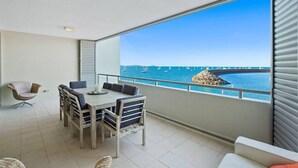 A balcony with a dining table and chairs, a wicker chair, and a view of the sea with boats.