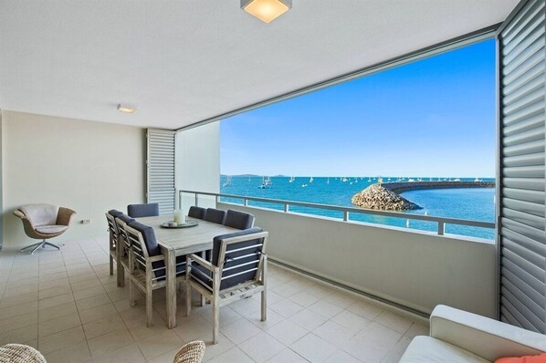 A balcony with a dining table and chairs, a wicker chair, and a view of the sea with boats.
