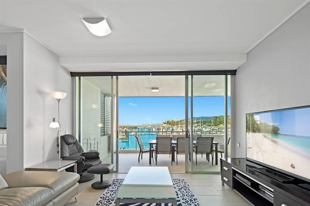 A modern living room with a large TV, comfortable seating, and a glass door leading to a balcony with a pool view.