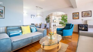 Una sala moderna con un sofá, sillones y una mesa de centro de vidrio. En el fondo, hay un comedor con una cocina visible.
