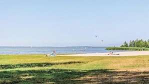 Ein Strand mit badenden und sonnenden Menschen, eine Rasenfläche im Vordergrund und Windkraftanlagen im Hintergrund.